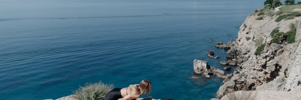 A couple performs a partner workout on a scenic cliffside, overlooking the ocean.